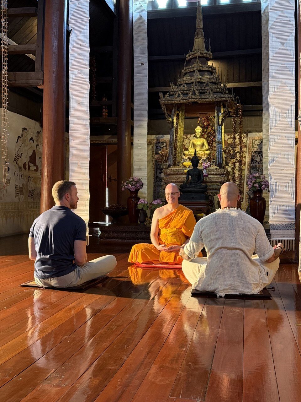 A monk of iMonastery leads Spirit Bright in seated meditation on the wooden floor of a temple hall before a golden Buddha at the Ancient City