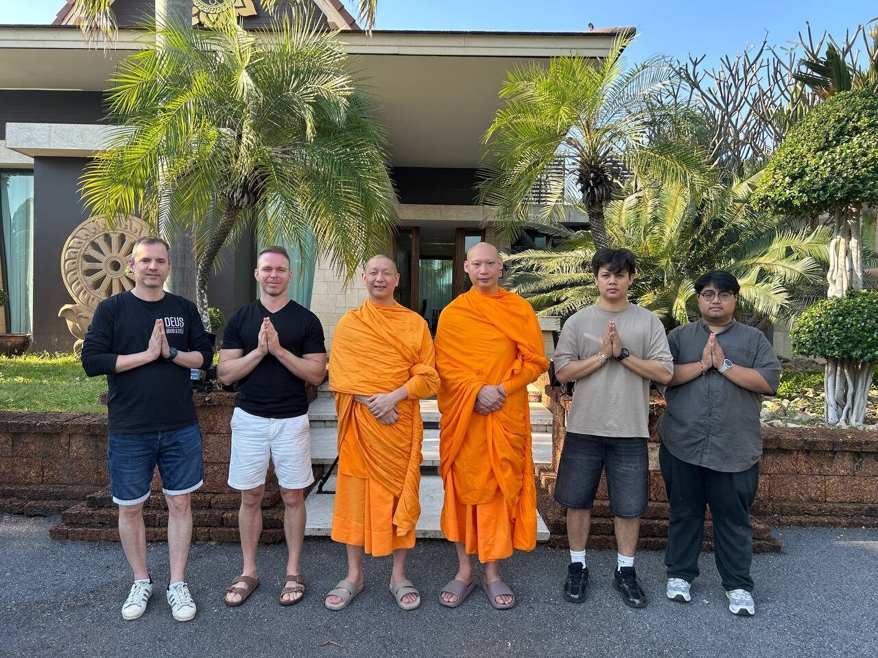 Group photo of the whole team in wai — two monks in orange robes at the centre, the film crew and Spirit Bright team either side, in front of the retreat house framed by palms.